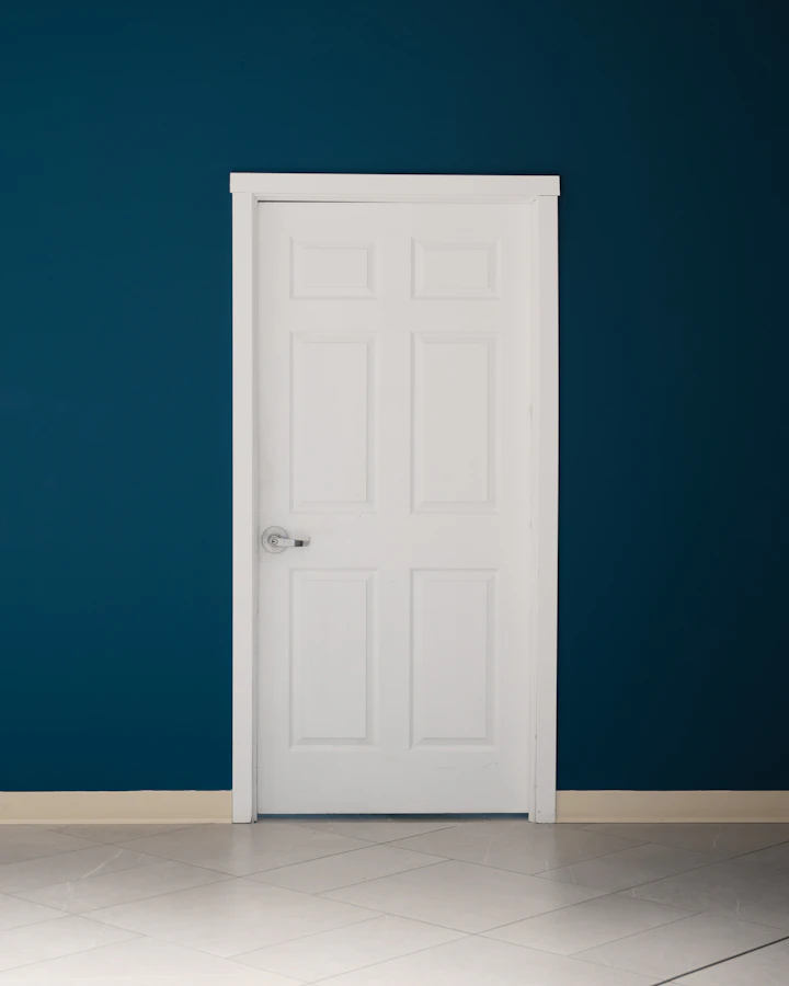 Porta branca com guarnições e batente destacados em parede de tom escuro (imagem ilustrativa)